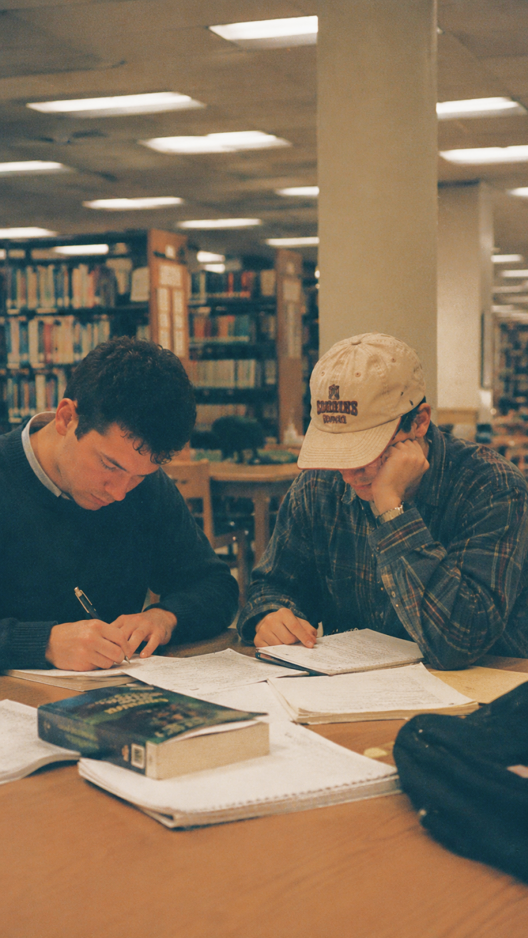 Two students writing notes together at a library table.